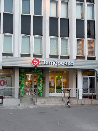 The image shows the exterior of a "Pyaterochka" (Пятёрочка) grocery store in Moskovskiy, Russia. The building has a modern facade with a repeating pattern of windows. The Pyaterochka logo, a red circle with a white stylized "5" and a green leaf, is prominently displayed on the building's vertical signage, which also spells out "Пятёрочка" in white letters. The entrance to the store is decorated with green and red balloons. Through the glass doors, shelves stocked with products are visible. To the right of the entrance, a purple electric scooter is parked near a railing. The area in front of the store appears to be a sidewalk and paved ground, with some debris scattered on the ground. The lighting suggests it is daytime. Another sign with the word "НИШ" and a red circle with a white "0" is visible below the main store entrance.