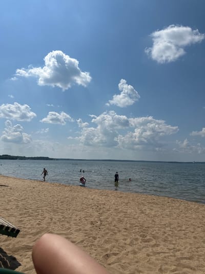 The image depicts a beach scene under a bright blue sky dotted with fluffy white clouds. The perspective is from the beach looking towards the water, with the camera rotated 90 degrees clockwise. The foreground is dominated by golden sand, with some debris scattered on its surface. A portion of a person's arm is visible in the lower right corner, suggesting the photo was taken by someone on the beach.
In the water, several people are visible. Some are wading near the shore, while others appear to be further out. One person in the middle ground is wearing a red top and blue shorts, and another person in the foreground is wearing dark shorts. There are also a couple of small red buoys in the water. The water itself appears calm and is a moderate blue color. In the distance, a faint treeline or shoreline is visible, suggesting a large body of water.
The overall atmosphere is one of a sunny day at the beach, likely during the summer. The weather appears pleasant with a clear sky and a few clouds, indicating a fair day. The scene suggests a leisurely activity, with people enjoying the water and the beach. The lighting indicates it is daytime, likely mid-morning or afternoon due to the bright sun. There is no visible text in the image.