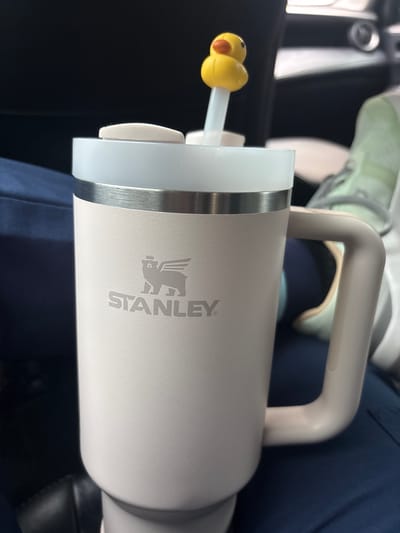 A light beige Stanley tumbler with a handle sits in what appears to be a car. The cup has a straw with a yellow duck attached to the end. The background includes a car door panel and an unidentified person wearing blue clothing and light-colored shoes. The scene is generic and does not provide information that reveals its location as Medellín, Colombia.
