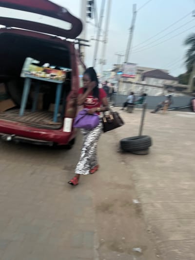 This media file captures a daytime street scene in Ablekuma, Ghana, rendered with a noticeable blur and rotated 90 degrees clockwise. The primary subject is a woman standing on a paved surface next to a parked red vehicle, which appears to be a car with its trunk open.

The woman has dark hair and is wearing a red top with an indiscernible white logo or design, and a distinctive shimmering skirt or trousers with a black and silver pattern. She has red open-toed shoes or sandals. She carries a dark brown handbag over her shoulder and a purple bag in her left hand. Her right hand is raised towards her face or ear.

The red vehicle, possibly a sedan or pick-up, has its trunk open, revealing a small, light-colored table with items on it, and what appear to be blue plastic frames or chairs inside. This setup suggests a possible commercial activity or preparation for loading/unloading goods.

In the middle ground, on the right, a distinctive makeshift pole stands upright with a car tire attached horizontally, possibly serving as a barrier or unique street furniture. The background features blurred outlines of buildings, utility poles, and overhead power lines, indicative of an urban or semi-urban environment. The ground is a mix of concrete or asphalt, showing some wear and discolouration. The time of day appears to be bright, likely overcast or daylight. No distinct emotions are discernible due to the image's blurriness. Visible text on the woman's shirt, car label, and distant signs is illegible.