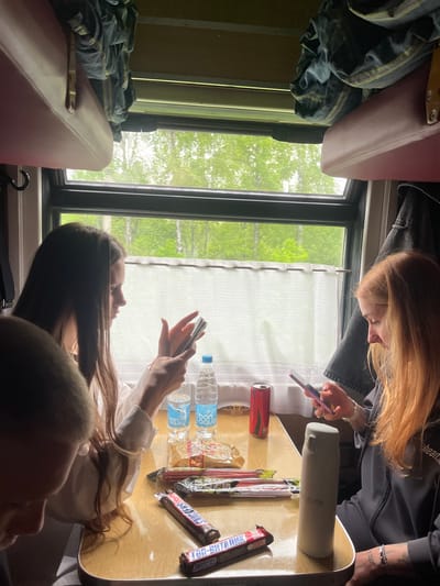 The image shows the interior of a train carriage, likely in transit. Two young women are visible, one with red hair and the other with dark hair. The red-haired woman is holding a phone. Both are seated at a table covered with snacks, including Snickers bars, bottles of water, and other packaged items. A can of soda is also visible. Outside the window, a green, wooded landscape is visible. The carriage has upper bunks with bedding. Based on the provided location, the train may be traveling through or near Красненскі сельскі Савет, Belarus.