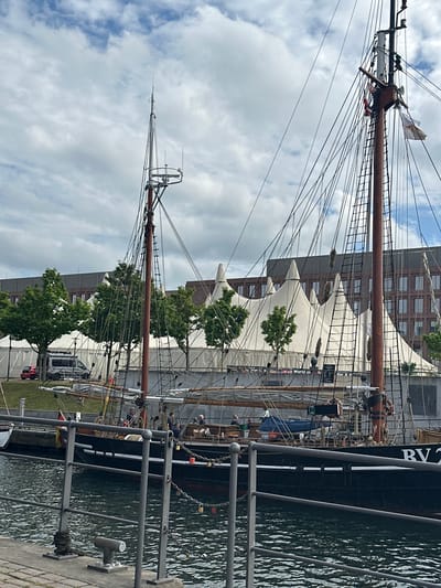 The image depicts a view of a tall ship docked in Kiel, Germany. The ship has tall wooden masts with rigging against a cloudy sky. Adjacent to the ship is a large white tent-like structure, with brick buildings visible in the background. A row of green trees adds a touch of nature, and a paved area with metal railings borders the water. People can be seen on and around the ship, suggesting possible activity or tourism. There are also cars and other vehicles parked nearby. The combination of maritime history, modern structures, and the presence of people suggests a tourist or public area in the city. The ship's hull displays the letters "BV 2".