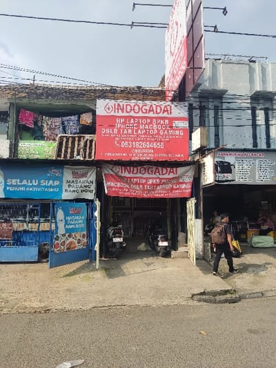 The image shows a storefront in Kecamatan Cimahi Selatan, Indonesia, located at latitude -6.899 and longitude 107.562. The storefront is a small shop with a red sign that reads "INDOGADAI HP LAPTOP BPKB IPHONE MACBOOK DSLR TAB LAPTOP GAMING CIBUREUI 083182604658." There are two motorcycles parked in front of the shop. The shop is located on a street with other businesses nearby. There is a man wearing a black shirt, black pants, and a backpack walking on the street. The man is walking away from the shop. There is a blue building behind the shop, and a large sign with the word "INDOGADAI" on it in the background. The environment is urban.