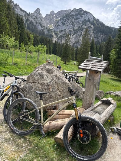The image shows a mountainous landscape, likely in or near Schwangau, Germany, judging by the presence of the Alps in the background. The foreground features several mountain bikes, resting near a wooden structure that appears to be a water fountain or trough. One person is visible standing or sitting near a large boulder, partially obscured by the bikes and the water fountain. The environment includes a grassy field with trees and a path. The scene is typical for an outdoor recreational area in the Alps, suggesting the bikes are used for mountain biking in the region.