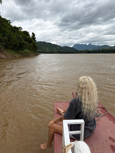 A woman with long, curly blonde hair is sitting on the edge of a red boat, facing away from the camera. She is wearing a dark grey t-shirt and light-colored shorts. Her left leg is extended, and her right leg is bent. She has a tattoo on her left arm. The boat is on a wide, murky brown river. In the background, lush green hills and mountains are visible under a cloudy sky. The scene appears to be in a rural area, possibly near Thinhong, Laos, given the natural landscape.