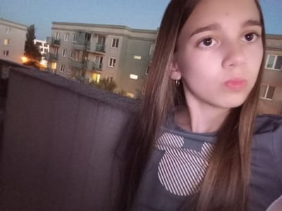 A young girl with long brown hair is taking a selfie. She has a slight pout on her lips and is looking towards the camera. She is wearing a grey t-shirt with a white Mickey Mouse print. She has a small hoop earring in her left ear.
The background shows a residential building with multiple stories and balconies. Lights are visible in some of the windows, suggesting it is evening or night. The sky is a dusky blue. To the left of the building, there appears to be a street light. A dark, textured surface, possibly a wall or roof, occupies the foreground and left side of the image.
The overall scene suggests a casual, personal moment captured outdoors. The time of day is likely dusk or early evening. The setting is urban or suburban, as indicated by the apartment building. The girl's expression is neutral, perhaps a little playful. There is no visible text in the image.