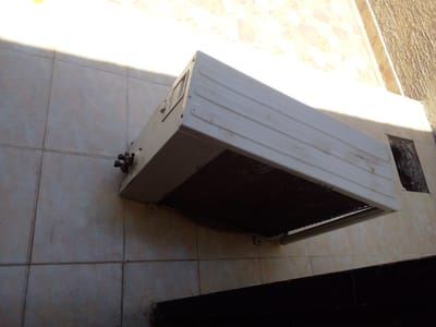 The image shows an outdoor unit of an air conditioning system mounted on a tiled wall. The unit is white and rectangular, with horizontal louvers on the front. There are pipes and connections visible on the left side of the unit. The wall is covered in light-colored square tiles, and there is a shadow cast across the wall from the left side, suggesting the sun is shining from the left. The background wall above the tiled section has a pattern with light and dark brown shapes. The overall scene appears to be an exterior wall of a building in a residential or commercial area. No people are visible in the image, and no text is present. The lighting suggests it is daytime. The scene is static, with the air conditioning unit simply mounted on the wall.
