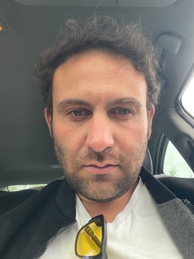 A man is pictured from a low angle, likely within a vehicle. He has dark hair, a beard, and light eyes. He is wearing a white t-shirt and a black jacket with a grey hood. Yellow-tinted sunglasses are visible near his chest. The interior of the vehicle is grey, with a sunroof visible at the top of the frame. The exact location in Armagh, United Kingdom cannot be determined from the image.