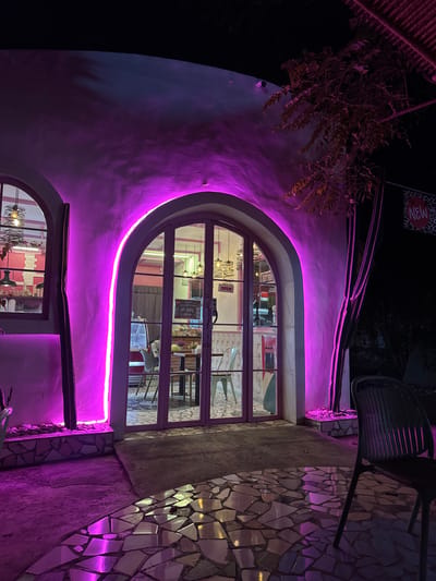 A night-time exterior shot showcases a building with an unusual, arched doorway and window illuminated by vibrant purple LED lighting. The architectural style features curved walls and a mosaic tile accent on the exterior, suggesting a whimsical or artistic establishment. Inside the arched window, tables and chairs are visible, along with what appears to be a bar area, indicating a restaurant, cafe, or similar venue. A sign with the word "NEW" in red and black lettering is partially visible at the top of the image, accompanied by a heart graphic. The overall atmosphere is festive and inviting due to the colorful lighting, despite the darkness of the night. The scene suggests the establishment is open for business, with interior lights on and furniture set up. The location context of Buleleng, Indonesia, adds a tropical or subtropical feel to the outdoor setting.