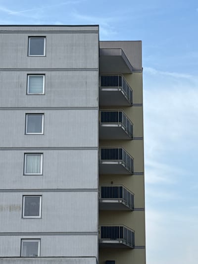 The image depicts the upper portion of a building, possibly located in the city of Grevenmacher, Luxembourg. The structure features a concrete facade with vertical columns and small, evenly spaced windows. Above the concrete section, there's a tan-colored section with five ventilation units, each protected by a slanted metal grate. The sky is visible in the background, showing a clear blue color with a few wispy clouds.