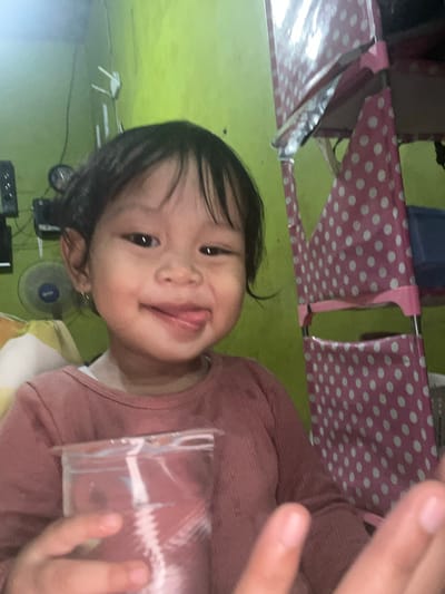 In an indoor setting, likely a home in Wedarijaksa, Indonesia, a young girl with dark hair and brown eyes is pictured. She is wearing a pink long-sleeved shirt and is holding a clear plastic cup filled with a pink beverage. The girl is sticking out her tongue playfully, with her right cheek slightly puffed out. Behind her, a green wall is visible, with a blue and white fan to her left. To her right, a pink shelf unit with white polka dots stands, partially obscuring a blue container. The lighting suggests a natural indoor environment.