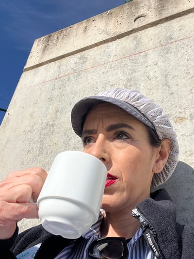 A person is holding a white mug in front of a concrete structure against a blue sky. The person is wearing a white and gray cap, a striped shirt, and a black jacket. Sunglasses are visible near the jacket. The photo appears to be taken in Mensdorf, Luxembourg, judging by the lighting and concrete construction style, which is typical of the region. The person is looking toward the camera with visible red lipstick.