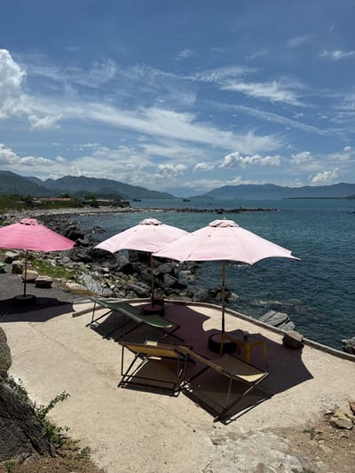 This is a photograph taken in Vĩnh Lương, Vietnam, showcasing a coastal scene with a rocky shoreline and calm turquoise water. In the foreground, a concrete pathway is lined with several pink beach umbrellas and empty lounge chairs, suggesting a recreational area. To the left, a large dark rock formation with sparse vegetation juts into the frame. The background features a hazy mountain range under a bright blue sky with scattered white clouds. The water appears relatively still, with a few small boats visible in the distance on the horizon. The overall impression is one of a serene, sunny day at a seaside location.