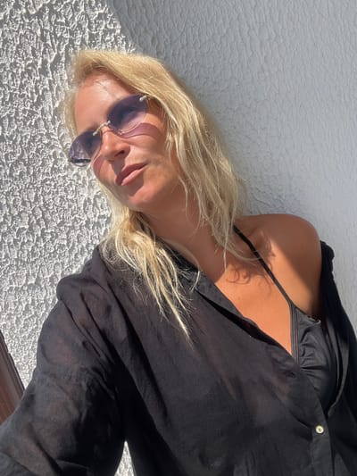 A blonde woman wearing a black unbuttoned shirt over a black bikini top and purple-tinted rimless sunglasses poses against a textured white wall. Sunlight casts a shadow across her face and illuminates her tanned skin. Her hair is loose and falls around her shoulders. The environment appears to be outdoors, possibly a balcony or patio, with a brown railing visible on the left edge of the frame. The scene is set in Giachrypsh, Abkhazia.