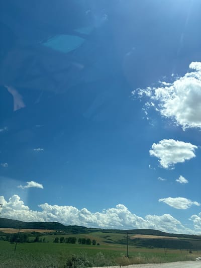 This is a wide-angle, eye-level shot of a rural landscape under a bright blue sky with scattered white clouds. The foreground shows a dirt road on the right and dry grass and plants on the left. Beyond that, there are rolling hills covered in green fields, with some areas appearing to be cultivated with crops. Patches of darker green indicate trees, and some areas show lighter brown or tan colors, possibly due to harvested fields. Several utility poles are visible throughout the middle ground. In the background, the hills continue, covered in a mix of trees and fields, under a vast sky dotted with puffy white clouds. The image appears to be taken from a moving vehicle, as suggested by the slight blur and reflections on what could be a car window in the upper left portion of the frame. The general scenery suggests a rural setting in or around Komarevo, Bulgaria, characterized by agricultural land and a clear, sunny day.