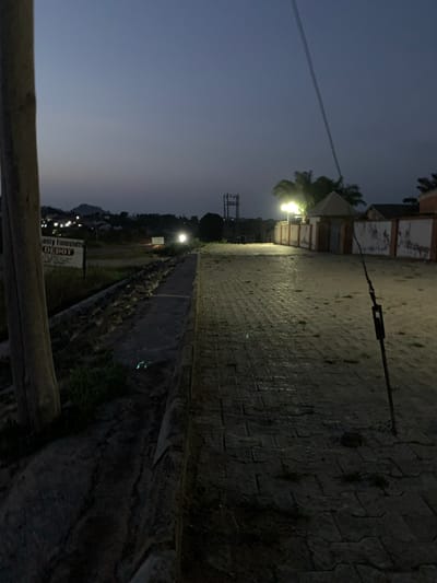 The image depicts a twilight scene on a paved walkway in Gbeleko, Nigeria. A wooden pole stands to the left of the frame, with a sign reading "nasty Foodstuffs DEPOT" visible behind it. The walkway is illuminated by streetlights, casting long shadows and highlighting the texture of the paving stones. A low stone wall runs along the left side of the walkway. In the background, buildings and a distant hill are visible, with scattered lights suggesting a developing urban area. A thin wire stretches diagonally across the upper right portion of the image. The overall impression is one of a quiet evening in a developing neighborhood.