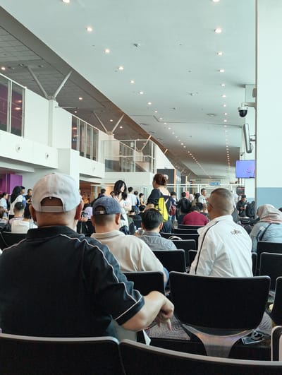 The image shows a waiting area inside Kuala Lumpur International Airport (KLIA) in Sepang, Malaysia. The area is brightly lit with overhead fluorescent lights and natural light from windows. Many passengers are seated in rows of black chairs. Some passengers are standing and walking. A television screen is mounted on a pillar. Signage in Malay is present. The architecture includes a modern design with high ceilings and support beams.