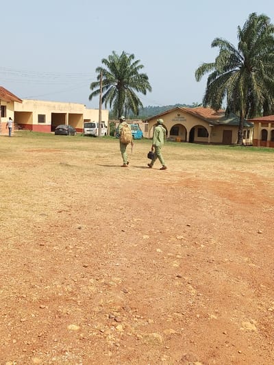 The image displays an open, unpaved area in Aramoko-Ekiti, Nigeria, under a clear, pale blue sky. The foreground consists of reddish-brown earth, interspersed with sparse dry, yellowish-green grass and small stones.

In the midground, two individuals wearing light-green uniforms, including matching caps, walk away from the viewer towards the right. The person on the left wears a light-colored backpack and holds a white rectangular object in their right hand. The person on the right carries a dark rectangular bag in their left hand. To the left of these individuals, another person in a blue shirt is visible near a building.

Vehicles parked in the midground include a dark grey sedan, two white vans, and a light-blue bus.

The background features several single-story buildings. On the left, a light-yellow building with a reddish-brown base and roof is visible. Centered in the background is a beige building with a dark brown corrugated roof and arched facade details, prominently displaying "JULIUS HOUSE" on a sign above the arches. A section of a similar building is visible to the far right. Several tall palm trees with green fronds are scattered across the scene, and a distant, tree-covered hill forms the horizon. The scene is brightly illuminated by direct sunlight.