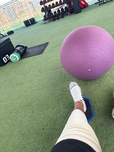 A purple exercise ball sits on a green floor, alongside a black mat and a weight bag labeled "15 KG". A leg wearing a white sock with black stripes and a compression sleeve is positioned with the foot on a blue and black sliding disc. A rack of dumbbells is visible in the background near a window.