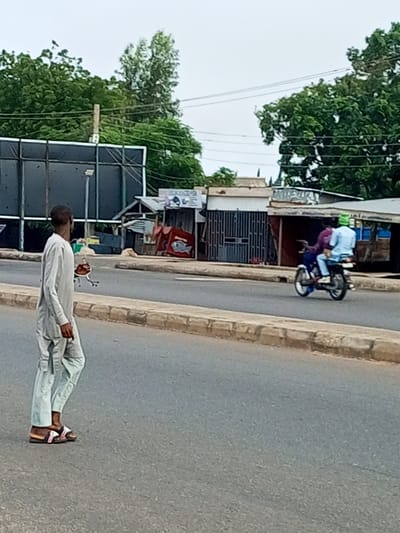 In Garko, Nigeria, a man in a light-colored traditional outfit walks on a paved road next to a curb. He is looking towards the left. In the background, two people are riding on a motorcycle. The person in front is wearing a purple shirt and the person behind is wearing a light blue shirt and a green cap. They are traveling on the same road as the pedestrian. Behind the road, there are several small buildings with corrugated metal roofs and some foliage. A large dark billboard is visible on the left side of the image. The sky is overcast.