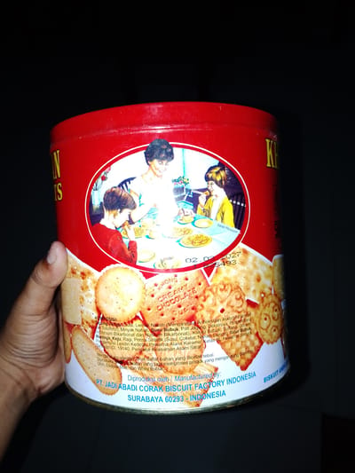 A hand is holding a red and white tin can with a picture of a family on it. The tin can is partially obscured by the hand and background. The image depicts three people, two children and one adult, gathered around a table. The adult is serving food to the children. The background shows a kitchen setting with cabinets and a window. The tin can also has text printed on it in white and yellow font. Part of the text reads "PT. JADI ABADI CORAK BISCUIT FACTORY INDONESIA SURABAYA 60293 - INDONESIA". The location is Sendangrejo, Indonesia.