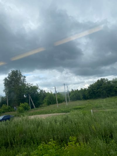 The image depicts a view of a rural landscape in the city of Nepeino, Russia, under a cloudy sky. In the foreground, dense green grass and vegetation are prominent. A dirt road runs horizontally across the middle ground. To the left of the road, a dark blue car is partially visible, parked or moving slowly. A large tree stands to the left of the road, with several utility poles and power lines extending across the scene. The background is filled with a dense line of green trees, suggesting a forested or heavily vegetated area. The sky is overcast with heavy gray clouds, with a faint horizontal light streak across the upper portion of the image, possibly a reflection or a light source. No people are clearly discernible in the image.