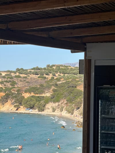 The image captures a view from a covered outdoor area, likely a restaurant or cafe, overlooking a beach and the Aegean Sea. The foreground is dominated by the wooden beams and ceiling of the structure, with a sliver of a refrigerator visible on the right. Beyond this, a vibrant turquoise sea with gentle waves stretches towards a sun-drenched beach. The beach is populated with numerous sunbathers and a few people wading in the water. Behind the beach, a rugged, hilly landscape covered in green scrub and scattered trees rises under a clear blue sky. The location appears to be Agios Nikolaos Municipal Unit, Greece, characterized by its coastal scenery and Mediterranean vegetation.