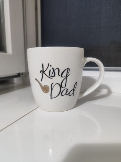 A white ceramic mug sits on a white tiled surface. The mug has a black script design that reads "King Dad" above a small illustration of a pipe. To the left of the mug is a white window frame, and behind the mug is a dark, textured surface, possibly a wall or screen. The overall scene is well-lit, with a shadow cast by the mug on the tiled surface. There are no people present in the image. The location is identified as Nerubaiske, Ukraine.