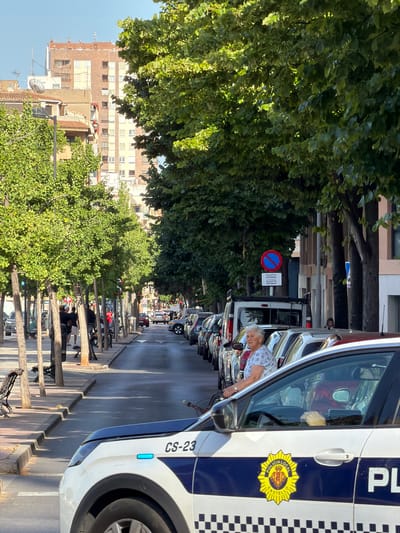 This is a vertical, eye-level shot of a street in Castelló de la Plana, Spain, likely taken in the late afternoon due to the long shadows. The scene is lined with trees and buildings, with parked cars and a police vehicle in the foreground.

The main subjects are the police vehicle and a woman leaning into its open driver's side window. The police car is white with blue and checkered markings, and the text "POLICIA LOCAL CASTELLÓ" is visible on its side, along with a circular emblem. The license plate reads "CS-23". The woman, with gray hair, is wearing a light-colored patterned shirt. She appears to be interacting with someone inside the police car, possibly an officer.

In the background, more parked cars are visible, along with bicycles and motorcycles parked along the sidewalk. The buildings lining the street are a mix of older brick and more modern facades. A circular red and blue traffic sign with text is also visible on a pole. The weather appears to be sunny and clear, with dappled sunlight filtering through the trees. The overall atmosphere seems to be that of a normal day in a Spanish city.