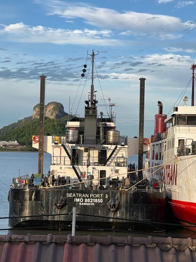 This is a vertically oriented photograph of two ships docked at a pier. The ship in the foreground is black with white text that reads "SEATRAN PORT 3" and "IMO 8621850 BANGKOK". A portion of a larger white ship with red accents is visible behind it. The ships are docked next to a brown, corrugated structure that appears to be part of the pier or a building on the pier. In the background, a lush green, rocky island rises from the calm blue water, and a hazy sky with white clouds is visible. The lighting suggests it is daytime, possibly late afternoon due to the angle of the sun and the shadows. The scene appears to be a harbor or a port in a tropical location, with the island in the background hinting at a Southeast Asian setting, potentially Thailand, given the text indicating Bangkok. There are no people visible in the image. The overall mood is serene and industrial, highlighting the maritime activity.