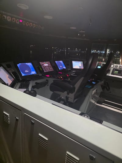 The image depicts the bridge of a ship at night, likely docked in the city of Brindisi, Italy. The dimly lit interior features a complex control console with multiple screens displaying navigational information and radar data. Two captain's chairs are positioned at the console, one of which is occupied by a person whose back is to the camera. Various buttons, switches, and a steering wheel are visible on the console. Outside the windows, the lights of a city and port are faintly visible against the dark night sky. The overall scene suggests a functional and operational vessel's command center.