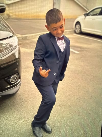An image captures a young boy dressed in a navy blue suit, white shirt, and a burgundy bow tie, standing outdoors on an asphalt surface. He is positioned to the left of the frame, facing the viewer with his body angled slightly to his right. The boy's right hand is raised, with his thumb and index finger extended in a gesture that resembles the "rock on" sign. His facial expression is a playful grimace, with his eyebrows furrowed and his eyes squinted.

To the boy's left, the front of a black car is partially visible. The car's headlight and bumper are distinct features. Further in the background, a white car is parked, and a light-colored building with a textured facade is also present. The overall environment suggests an outdoor urban setting. The image appears to be taken during daylight, with natural lighting illuminating the scene. The absence of any text or specific landmarks within the image makes it difficult to ascertain the exact location within Artik, Armenia, or any other city.