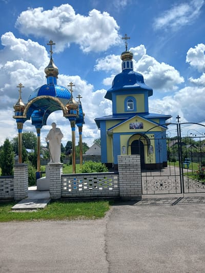 In Doslidnytske, Ukraine, a bright blue and yellow church stands under a cloudy sky. To the left, a decorative blue and gold canopy shelters a white statue of a robed figure holding a staff and a book. The canopy is supported by golden columns and topped with smaller golden domes. A white brick wall with decorative cutouts separates the churchyard from a paved area. A black wrought-iron gate with ornate scrollwork guards the entrance to the church property. In the background, green trees and houses are visible. The overall scene depicts a serene religious site on a partly cloudy day.