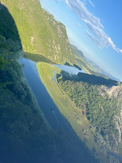 The image shows a wide, winding river flowing through a valley surrounded by steep, tree-covered mountains. The water is dark blue and reflects the sky and surrounding landscape. A thin band of green vegetation lines the banks of the river, and beyond that, the slopes of the mountains rise dramatically, covered in dense green foliage. The sky is a clear blue with scattered white clouds. The scene is shot from a high vantage point, looking down and across the valley. A small motorboat is visible on the river, leaving a wake behind it. The lighting suggests it is either early morning or late afternoon, with a strong shadow cast from a mountain on the left side of the image. The image appears to be from the Skadar Lake National Park, near Rvaši, Montenegro.