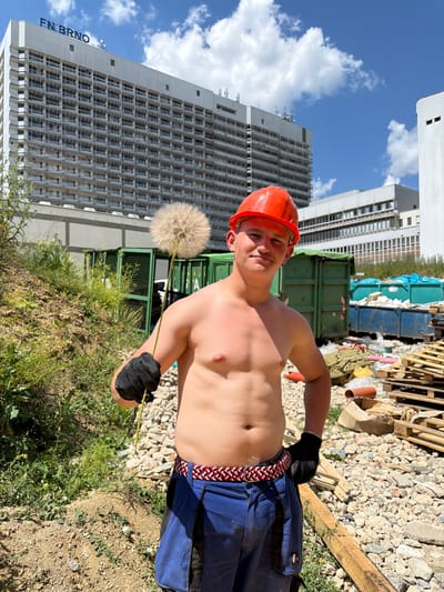 A bare-chested young man wearing a bright orange construction helmet and blue work pants stands in what appears to be a construction or demolition site. He is holding a large dandelion seed head in his gloved left hand. Behind him, large green and blue dumpsters are visible, filled with debris and construction materials. Pallets and wooden planks are also scattered around the site. In the background, a massive, multi-story white building with many windows dominates the skyline. The building has "FN BRNO" written in blue letters across its top. The sky is blue with scattered white clouds. The ground is covered in dirt, gravel, and rubble. The overall impression is that of a construction site in the city of Brno, Czechia, possibly near the FN Brno hospital.