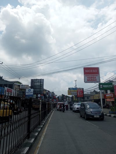 The image shows a street scene in an unidentified city. The sky is overcast with large, puffy clouds. Electrical wires crisscross overhead. The left side of the street is lined with a black metal fence and a variety of businesses and signs. Prominent signs advertise "JELITA FASHION," "PUSAT GADAI INDONESIA," "NICESO" with the phrase "SEMUA BARANG HARGA MURAH," "BINGYU" ice cream and coffee, and "MOMOYO." The street has light traffic consisting of cars and motorcycles. A gray minivan is in the foreground, with license plate "D 1128 NH." Yellow vans are visible behind the fence. There are a few people walking along the sidewalks near the businesses. The overall impression is of a bustling commercial district.