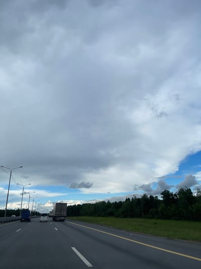 The image shows a view from a vehicle driving on a highway near Бавыкино, Russia. The sky is dominated by heavy cloud cover, with patches of blue visible. To the right of the road, there is a line of trees bordering a grassy area. The road is asphalt with white lane markers and a yellow line on the right edge. A line of streetlights runs along the side of the highway. Several vehicles, including a semi-truck and smaller cars, are visible on the road ahead.