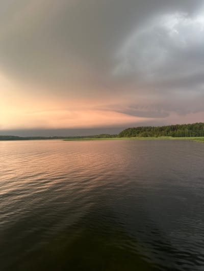 This is a landscape photograph of a wide body of water, likely a lake, under a dramatic sky. The water is dark and reflects the colors of the sky, with subtle ripples on its surface. The shoreline is lined with a dense forest of green trees, with some taller reeds or grasses visible at the water's edge. The sky is a dynamic mix of colors, with soft pinks and oranges on the left, transitioning to darker grays and blues on the right, suggesting a storm or sunset. There are no people or man-made structures visible in the image, creating a natural and serene scene. The location is identified as Слабодкаўскі сельскі Савет, Belarus.
