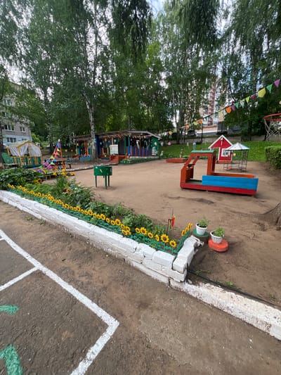 The image depicts a vibrant and colorful playground for children in Советско-Никольское, Russia. The playground is situated outdoors, surrounded by lush green trees and with residential buildings visible in the background. The ground is primarily covered with sand, a common surface for playgrounds.

Dominating the scene is a red and blue sandbox structure, designed to resemble a boat or train. Adjacent to it is a small red house with a white roof and blue trim, and a colorful canopy structure with a slide. Colorful flags are strung across the playground, adding to the festive atmosphere. A white brick border, adorned with artificial sunflowers, lines a section of the playground. Two small potted plants sit on decorative orange stands near the sunflowers.

In the foreground, there are white painted lines on the ground, possibly indicating a game area or boundary. A green metal bin is also present in the middle ground. The playground appears to be well-maintained and equipped with various play structures and decorative elements, suggesting it is a dedicated area for children's recreation. The overall impression is one of a cheerful and inviting space for outdoor play.
