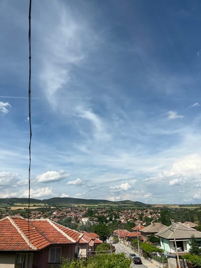 This is a daytime, high-angle, outdoor shot of a village in Popintsi, Bulgaria. The foreground is dominated by the terracotta tiled roofs of several houses. A dark wire, likely a power or telephone line, runs vertically down the left side of the frame, obscuring a portion of the sky and village.
Beyond the houses, the village sprawls across rolling hills. The buildings are predominantly low-rise, with red or grey tiled roofs and light-colored walls. Green trees and vegetation are interspersed throughout the village and the surrounding landscape. The background consists of green hills and fields, with a hazy, light-blue sky dotted with white, wispy clouds. A contrail is visible in the upper left quadrant of the sky. A paved road with parked cars is visible winding through the village. A few figures are subtly present on the road, though not clearly discernible. The overall impression is one of a rural Bulgarian village on a partly cloudy day.