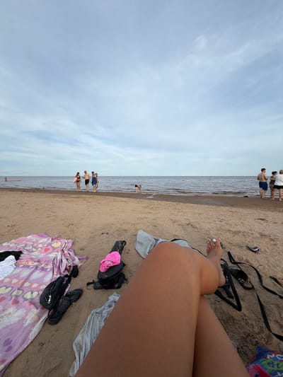 The image depicts a group of people on a sandy beach, with the ocean and sky in the background. The scene is oriented vertically, with the sky occupying the left side and the beach and water on the right.

In the foreground, a person's legs are visible, with their feet and toes painted white. Various objects are scattered on the sand around them, including a pink item of clothing, several bags, and a pair of black sandals. A patterned towel is also visible, laid out on the sand.

Further back, a group of people are standing near the water's edge. Some are in the shallow water, while others are on the sand. They appear to be dressed in swimwear. In the distance, more people are visible in the water. The water itself is calm, and there are buoys or markers in the distance.

The sky is overcast with some wispy clouds. The overall lighting suggests it is daytime, possibly late afternoon or early evening, given the diffused light. The presence of beach attire and the bodies of water suggests a recreational setting, likely a seaside location. The overall mood seems relaxed and casual. There is no visible text in the image.