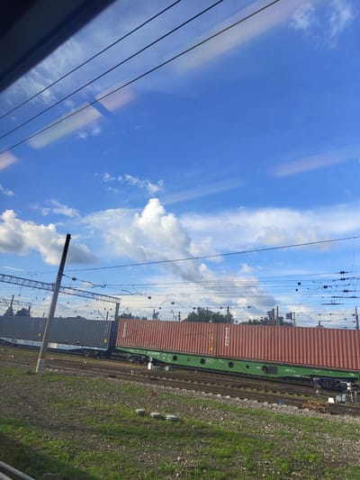 The image shows a freight train with two containers on a railway track. The train is positioned on a slight incline, with the front of the train in the foreground and the rear receding into the distance. The first container is dark grey and appears to be of standard size. The second container, which is closer to the foreground, is a reddish-brown color and is also a standard shipping container. The train cars are green. The railway tracks are laid on a bed of gravel and ballast, with patches of green grass growing in between. In the background, there is a line of trees and a clear blue sky with scattered white clouds. Overhead, there are power lines and supporting structures for the railway electrification system. The image appears to have been taken from a moving vehicle, possibly a train, looking out of a window, as evidenced by the reflections and the slight blur in some areas. The scene is likely a railway yard or a section of track near a logistics hub. No people are visible in the image.