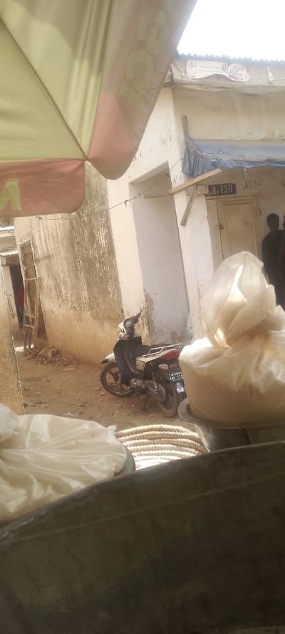 The image depicts an outdoor urban scene, likely a market or residential street in Nigeria, under bright daytime illumination. The foreground is partially obscured by commercial elements: the underside of a two-tone (pale yellow and reddish-pink) umbrella with "FOR GREAT TASTE PICK MAGGI STAR" and a star logo faintly visible, and two transparent plastic bags containing light-colored granular material resting in a dark, shallow pan. A woven, light brown basket or mat is visible beneath these items. The very bottom of the frame is obscured by a dark, flat surface.
In the midground, a dirt or unpaved pathway extends into the scene. A white and black scooter/motorcycle with license plate "KWS-350" is parked on this path, facing left. Buildings with weathered, light-colored walls, showing signs of dirt and peeling plaster, flank the pathway. The building on the right features a light-colored wooden door with "JA. 1371" and "ASSO" marked on the wall above it. A dark blue tarp is draped from the roofline, secured by a wooden plank. The building on the left has a wooden structure, resembling a ladder or support, leaning against its wall.
Two indistinct human figures are partially visible in the far background on the right, near the building entrance, one attired in dark clothing. The sky above is bright and hazy. The specific proximity to Muhammadu Dikko Stadium in Nigeria cannot be determined from the visual evidence.