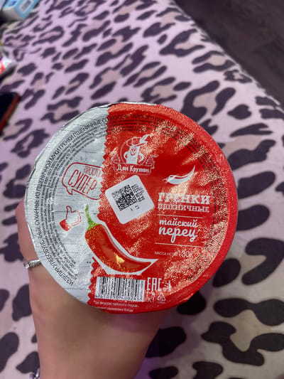 A person's hand with a ring is holding a round container with a red lid. The lid has text and images, including a chili pepper and a QR code. The background is a leopard-print fabric. The container appears to be food, possibly a snack. There is no visible information indicating the location in Borisova Griva Massiv, Russia.