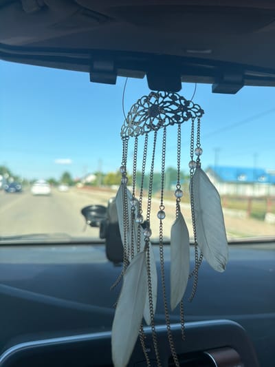 An ornate, bronze-colored dreamcatcher with white feathers hangs from the rearview mirror inside a car. The dreamcatcher is intricately designed with floral patterns and chains adorned with small, clear beads. The car's interior is visible in the foreground, with a dark dashboard. Through the windshield, a bright blue sky with a few clouds is visible. The exterior scene shows a paved road with several cars driving in the distance, suggesting a suburban or semi-urban environment. Houses and trees line the roadside, and the overall impression is of a sunny day. The scene is located in the city of Duliby, Ukraine.