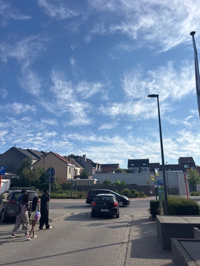 The image depicts a street scene in Strombeek-Bever, Belgium, on a clear day with wispy clouds. In the foreground, a grey tiled walkway is visible, with a dark grey, rectangular planter box to the right. Several people are walking on the walkway towards the left of the frame, including two women and two men. One woman is wearing a patterned top and loose pants, and the other is wearing a light-colored dress. A man is wearing a black tracksuit. To the left of the people, a dark grey SUV is parked.
Further back, a street is visible with several cars. A black hatchback with license plate "1 THZ-152" is parked on the right side of the street. A black sedan is driving in the background. A tall, dark lamppost stands prominently in the middle ground to the right of the image, and another shorter lamppost is visible to its left. Various residential buildings with tiled roofs and some signage are present in the background, suggesting a suburban or mixed-use area. A blue parking sign with a wheelchair symbol is also visible on the left side of the image.