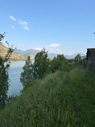 The image depicts a serene natural landscape in the vicinity of Anos, Russia. A wide, tranquil river occupies the left side of the frame, bordered by verdant, undulating hills in the background. The foreground is dominated by a grassy slope, with a narrow dirt path winding its way upwards. On the right side of the image, a rustic, dilapidated wooden fence and a dilapidated structure with a corrugated roof suggest the presence of a remote dwelling or an abandoned farmstead. Tall trees, including birch trees, are scattered throughout the scene, adding to the natural beauty. The sky is a clear, bright blue with a few scattered white clouds. The overall impression is one of peaceful isolation and rural simplicity.