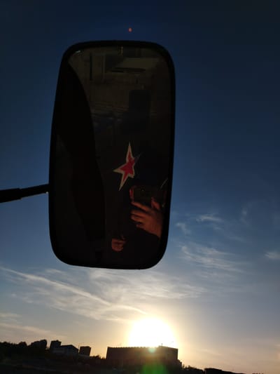 In Baikonur, Kazakhstan, a side-view mirror of a vehicle reflects the sky and a hand holding a smartphone. A red star emblem is visible on the clothing of the person in the reflection. The background of the mirror shows interior elements of the vehicle and a distant building. Outside the mirror, the sky is a gradient of deep blue to lighter hues, with wispy clouds illuminated by the setting sun. The horizon features a silhouette of urban buildings and trees against the bright sun.