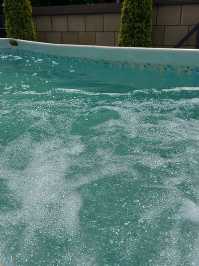 The image depicts a close-up view of a partially filled swimming pool, with clear, turquoise water and numerous small bubbles creating a frothy surface. The pool's edge is visible, featuring a light-colored coping with a decorative tiled border in shades of blue and white. In the background, a section of a stone wall with horizontal lines separates the pool area from a dark brown fence. Two tall, green, cone-shaped shrubs are positioned behind the pool, framing the scene. The lighting suggests it is daytime, with sunlight reflecting off the water's surface. The image does not contain any people or specific identifiable landmarks to pinpoint a location within Koroviia, Ukraine.
