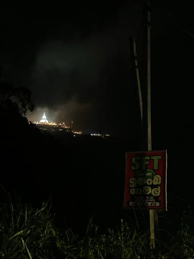 A night-time view in Kumbalwela, Sri Lanka, features a sign for "SFT Tea Factory" in Sinhala script attached to a pole. In the background, a white stupa is illuminated on a hill, surrounded by other lights marking the location of buildings or streets. The foreground is dark, with vegetation on the right side of the frame. Power lines are visible above the sign. The lighting is dim.